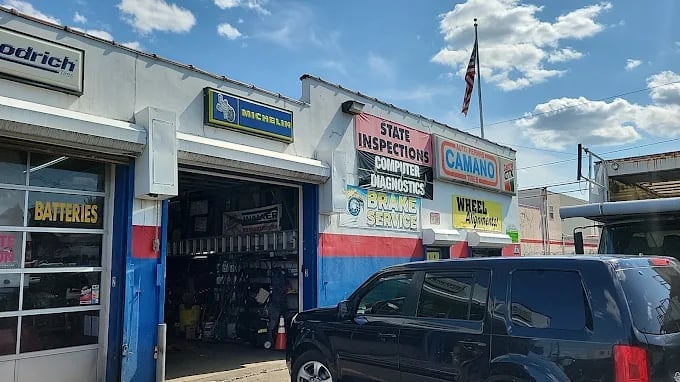 Auto repair shop storefront with blue and white exterior, multiple business signs including batteries and Camaro signage, parked dark SUV, and American flag